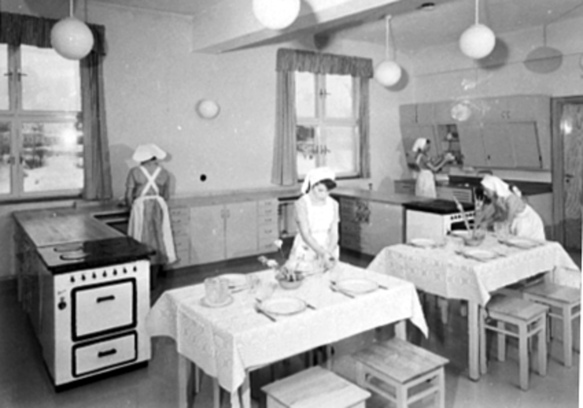 Anno Domkirkeodden, INTERIØR SKOLEKJØKKENET PÅ STORHAMAR SKOLE, FIRE JENTER MED FORKLÆR OG SKAUT, RYDDER OG DEKKER MIDDAGSBORD, FREDRIK MONSENS GATE 52.