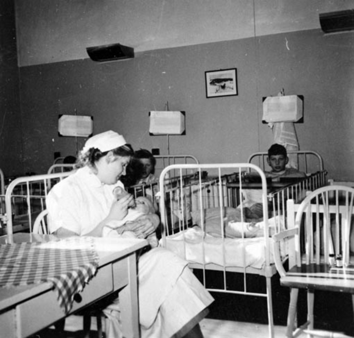 Anno Domkirkeodden, HAMAR RØDE KORS KLINIKK. BARNEPLEIER GIR SPEBARN MELK FRA TÅTEFLASKE. BARNEAVDELINGEN. BARN I SENGER,