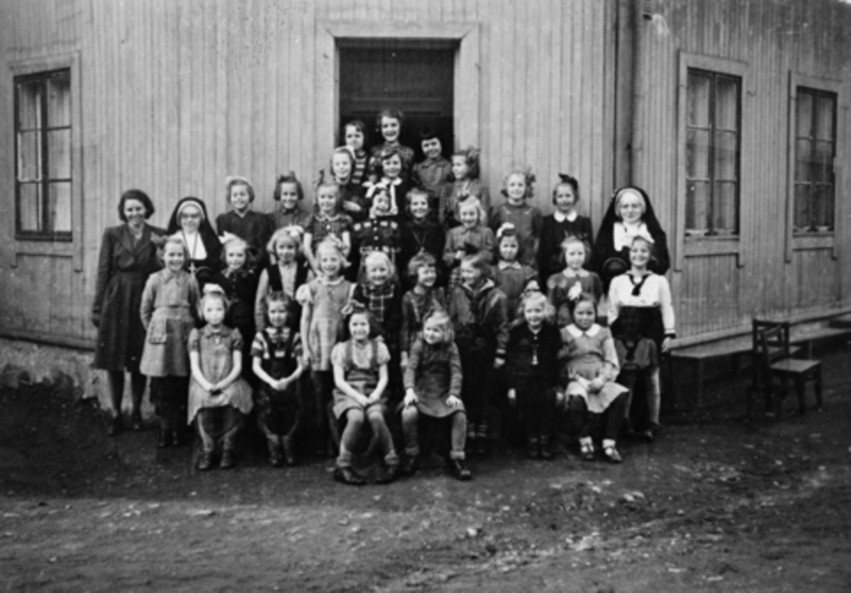 Anno Domkirkeodden, ST. TORFINN, KATOLSKE KIRKE HAMAR, GRUPPE JENTER OPPSTILT I REKKE UTENFOR KIRKEN, PYNTET I KJOLER SLØYFER I HÅRET, NONNE, UKJENT ÅR.
