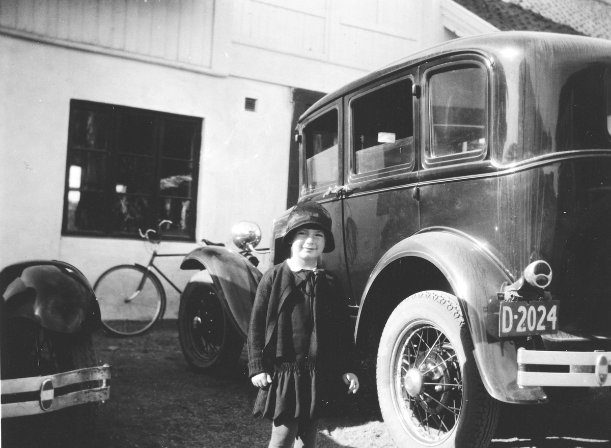Anno Domkirkeodden, Randi Andresen, gift Meyer. (1927-2009). Bil D-2024, Oplandske auto i Enggata, Hamar. D-2024 er et prøvenummer. Den røde bakgrunnsfarven fremtrer som mørk i s/hv. Formodentlig tilhørte nummeret Oplandske Auto. Bilen er en Ford modell A 1930-31. Til venstre støtfanger og forskjerm til en Ford A 1928…