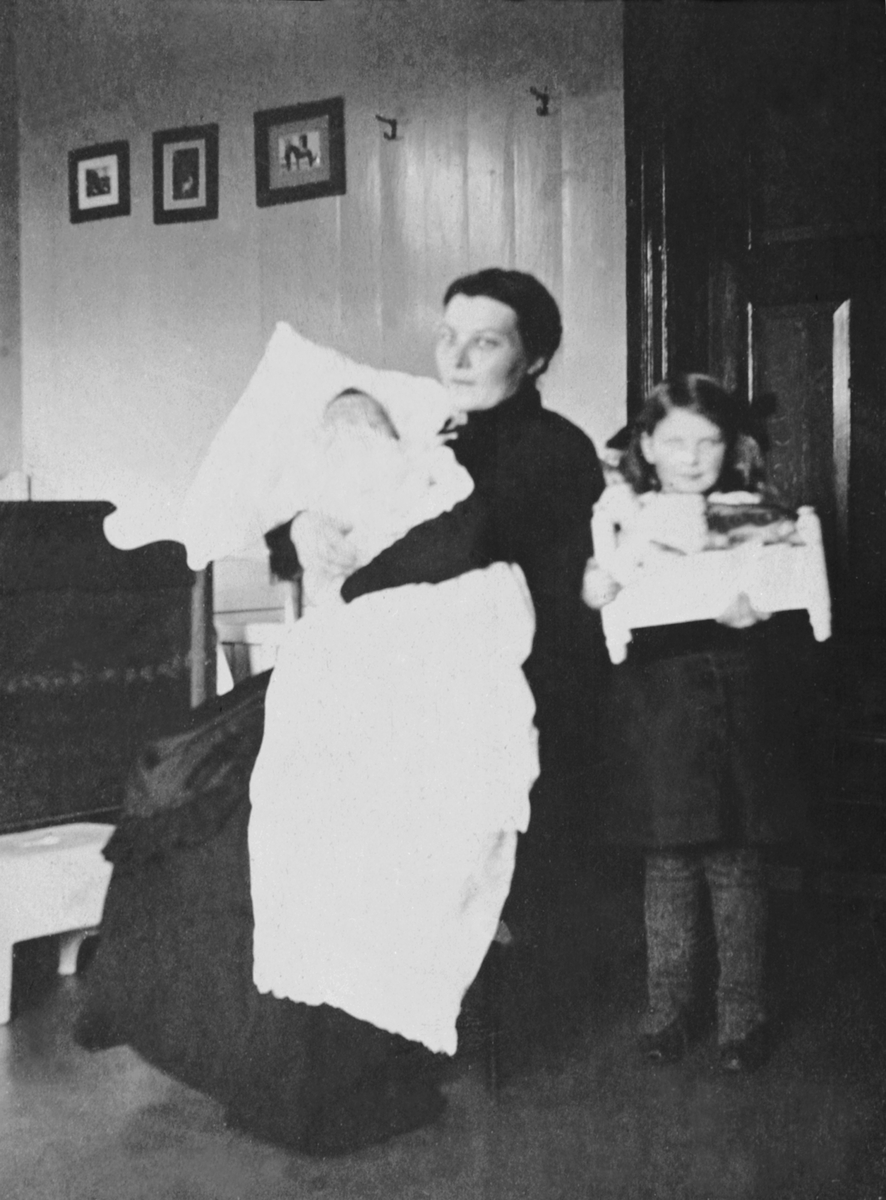 Anno Domkirkeodden, Interiør soverom på Mellom-Kise, Nes, Hedmark. Mor og barn. Anne Dorthea Haugen (1883-1965) med datteren Kristiane Lise Mathea (1914-1995) med dåpskjole på fanget. Anna Marie Haugen (1910-1992) ved siden av med dukkeseng. Dukkesenga var gave på 2 årsdagen.