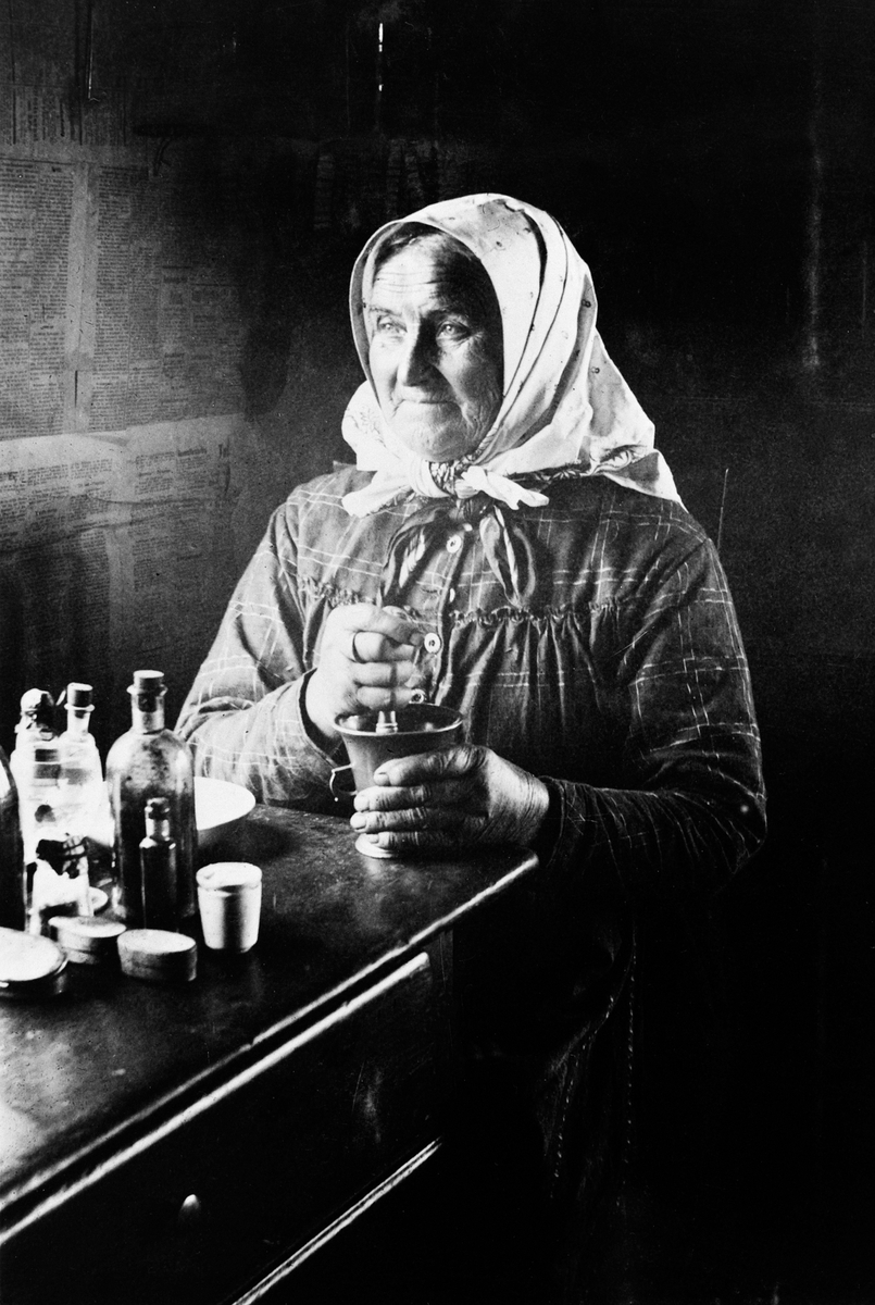 Anno Domkirkeodden, Portrett av eldre dame med skaut som sitter med morter ved bord. Kansje hun blander medisin, urter. Veggene er kledd med avispapir.