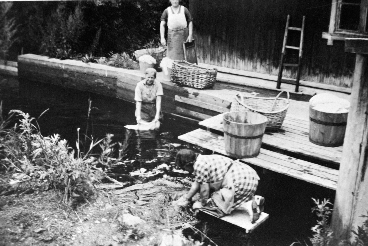 Anno Domkirkeodden, Vasking av klær foran K.Kolloens vaskehus på Finsdal, Brumunddal. Bryggerhus, kvernveita. Foran er Margrethe Kolloen, Laura Hansen. Bak er Julie Olsen. På bildet ser en at det er anlagt små brygger hvor man kunne ligge å skylle klær. Her er imidlertid vannstanden så lav at Laura Hansen har vasset u…