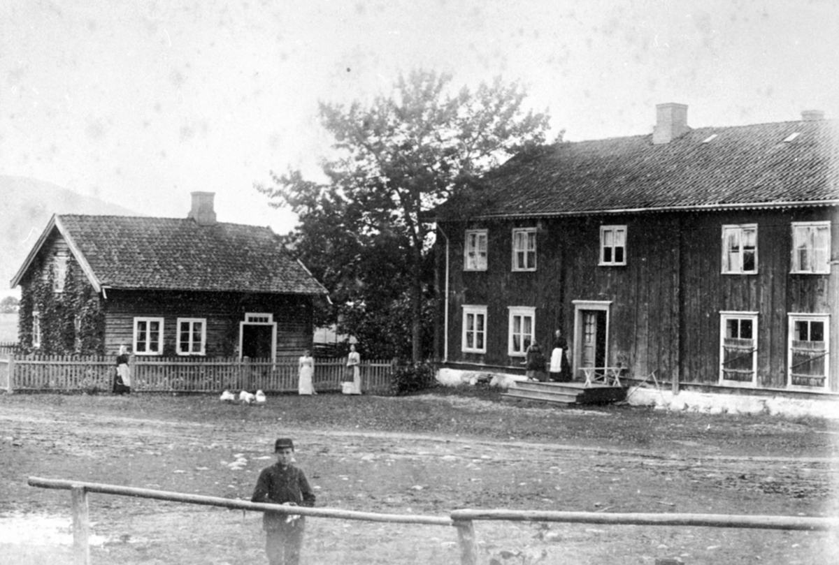 Anno Domkirkeodden, Eksteriør, gardsbruk, Kvernengen før flytting, inntun, folk på tunet, tidligere landhandel. Oline Jevanord med hvitt forkle på trappa. Foran Raustugua står Karen Julie Jevanord gift med baker Kristiansen. Brumunddal stasjon ligger her i dag. Den liile stua til venstre ble flyttet opp i Enga og står…