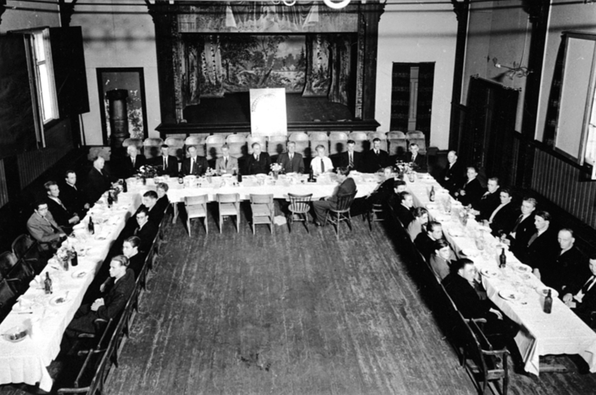 Anno Domkirkeodden, Festmiddag for ansatte på Globus Maskinfabrikk. Nils Ambli foran scena, interiør Framheim kino, Brumunddal. Dekket langbord.