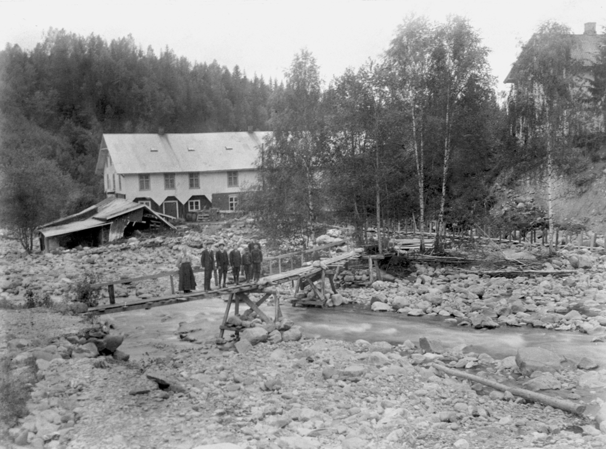 Anno Domkirkeodden, Brumunda, etter storflom, våren 1916. Bygingen i bakgrunnen er Brumunddal Ullvarefabrikk, tidligere Narud Uldspinneri. 7 ukjente personer på bru over elva.