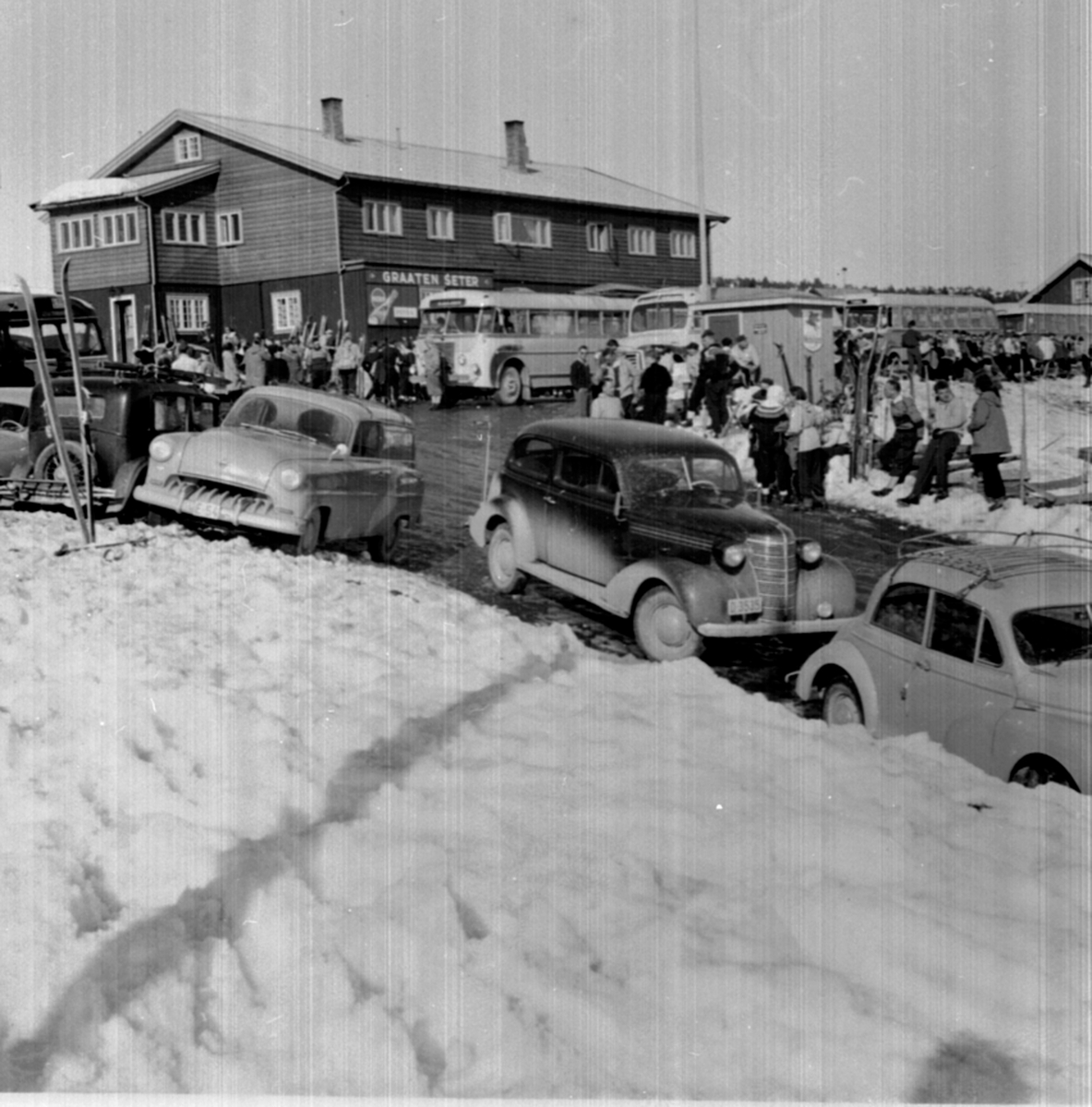 Anno Domkirkeodden, Sjusjøen, bussavgang fra Graaten til Hamar og Lillehammer. Mye folk, påsketrafikk. Bilene i forgrunn er fra venstre ukjent merke ca. 1932, E-54 Opel Rekord varebil 1953, Den mørke bilen, D-3535, er en Chevrolet 1938, Morris Minor.