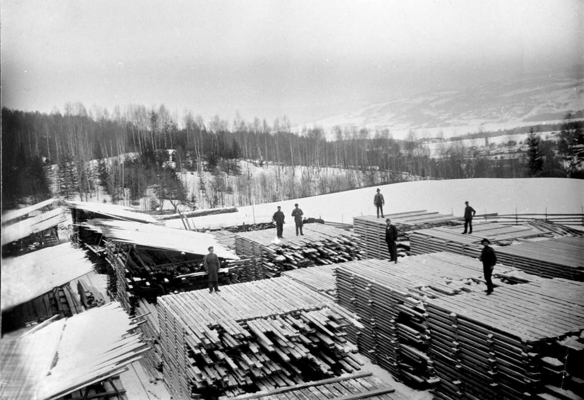 Anno Domkirkeodden, Oversikt over materialtomta på Moelven Brug. Lager av materialer som er stikklagt til tørk. Syv mann arbeider. Vinter.