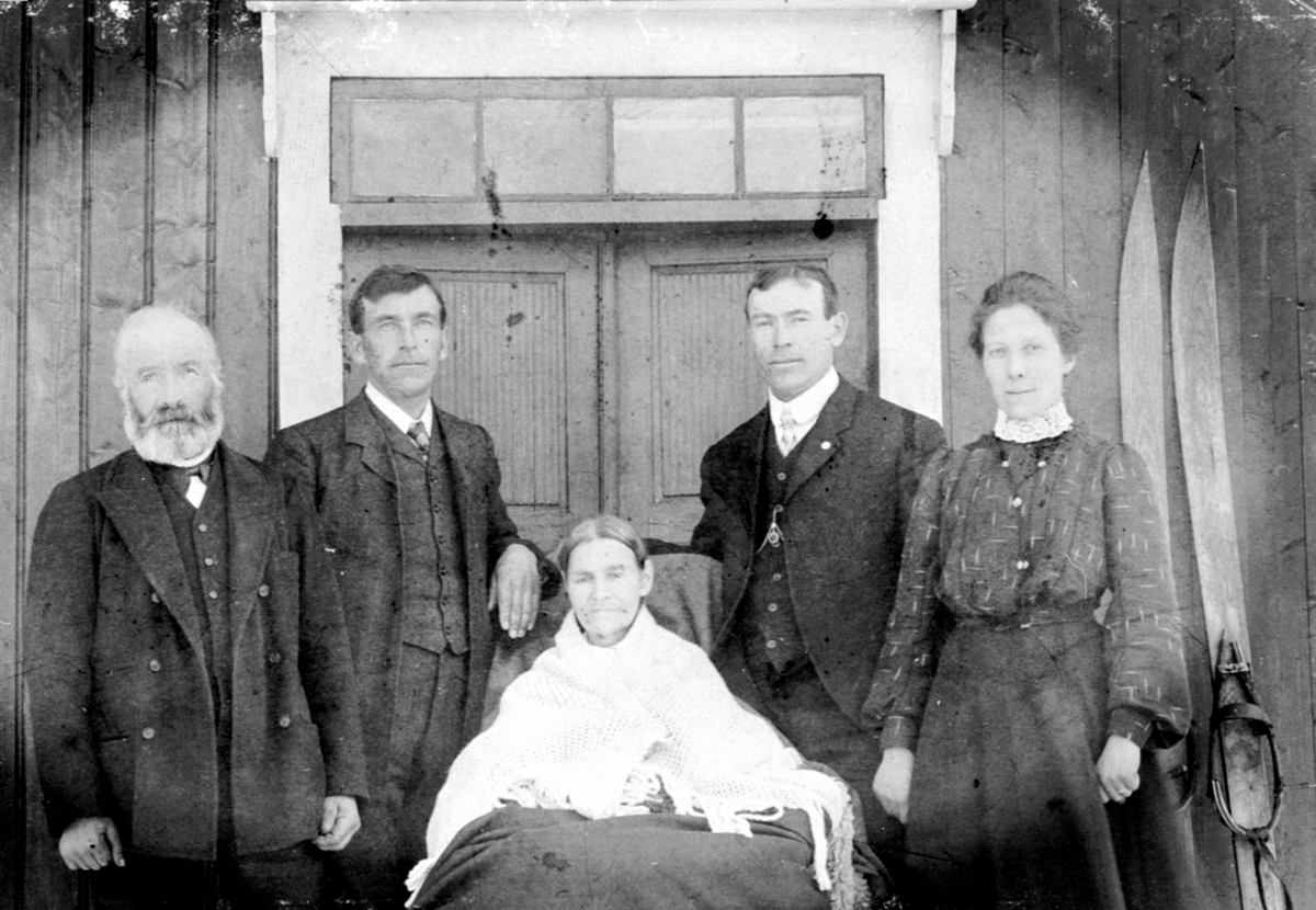 Anno Domkirkeodden, 5 personer foran huset i Vestgård, Helgøya. Fra venstre Ole Andersen Vestgård (1844-1908), Tonette Vestgård (1839-1923) sittende i midten og Trine Vestgård til høyre. 2 sønner som emigrerte til Amerika står bak. Et par gamle ski ved siden av døra.