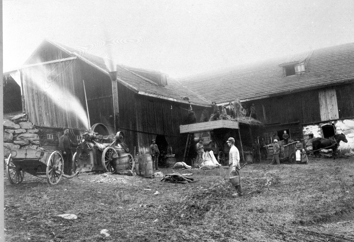 Anno Domkirkeodden, Biskoplien nedre, Nes, Hedmark. Trøsking med trøskemaskin. Trøskeverket drives av lokomobil. Nils Lien ved høykjerra, Per Arnseth foran med bøtte. Lise Godager holder sin nevø Even (1903-1905) i handa.