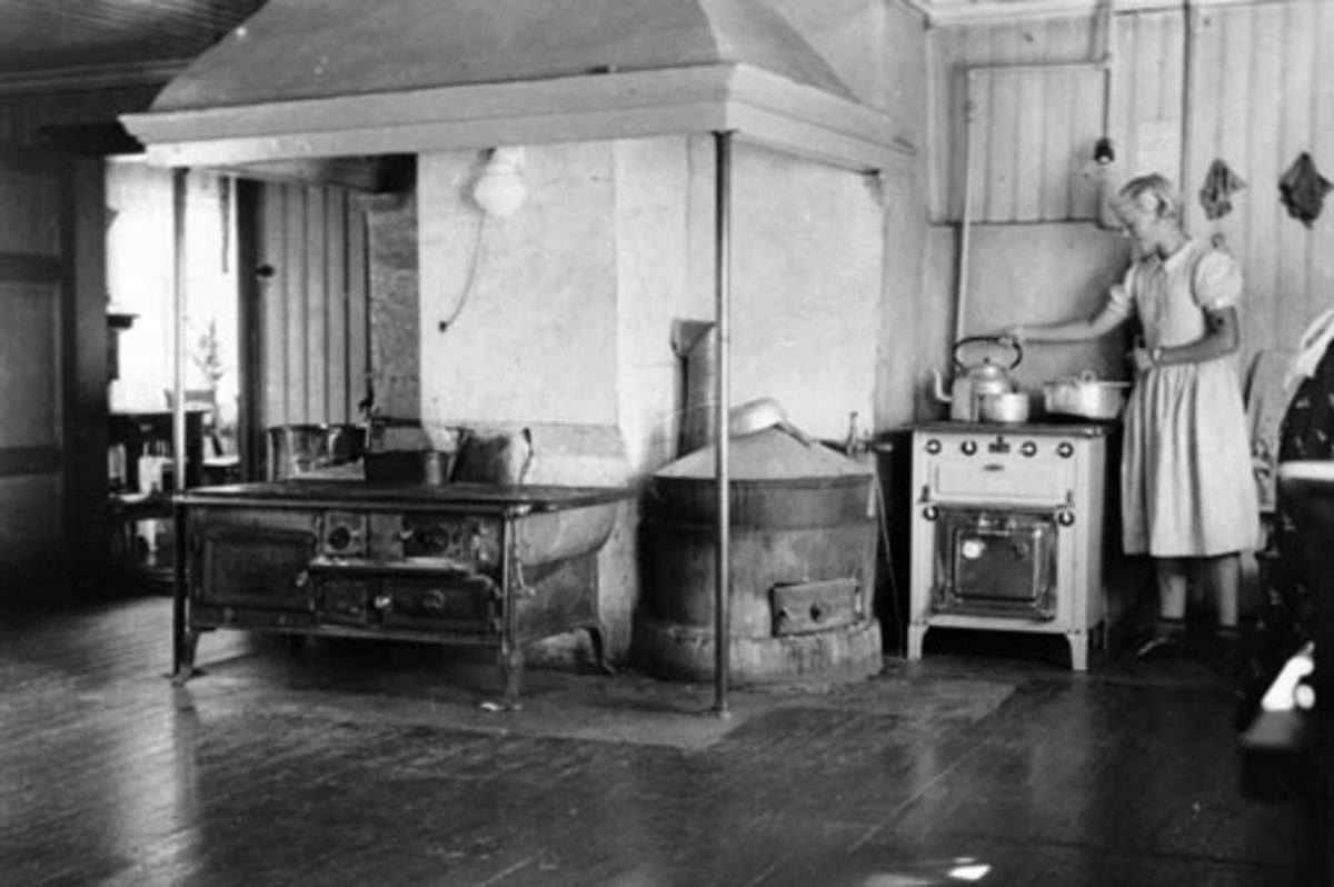 Anno Domkirkeodden, Interiør fra kjøkkenet på Ousdal nordre, Nes, Hedmark. Peis med innsatt vedkonfyr, bryggpanne, elektrisk komfyr. Margit Leich Ousdal f.1923 ved kaffekjelen.