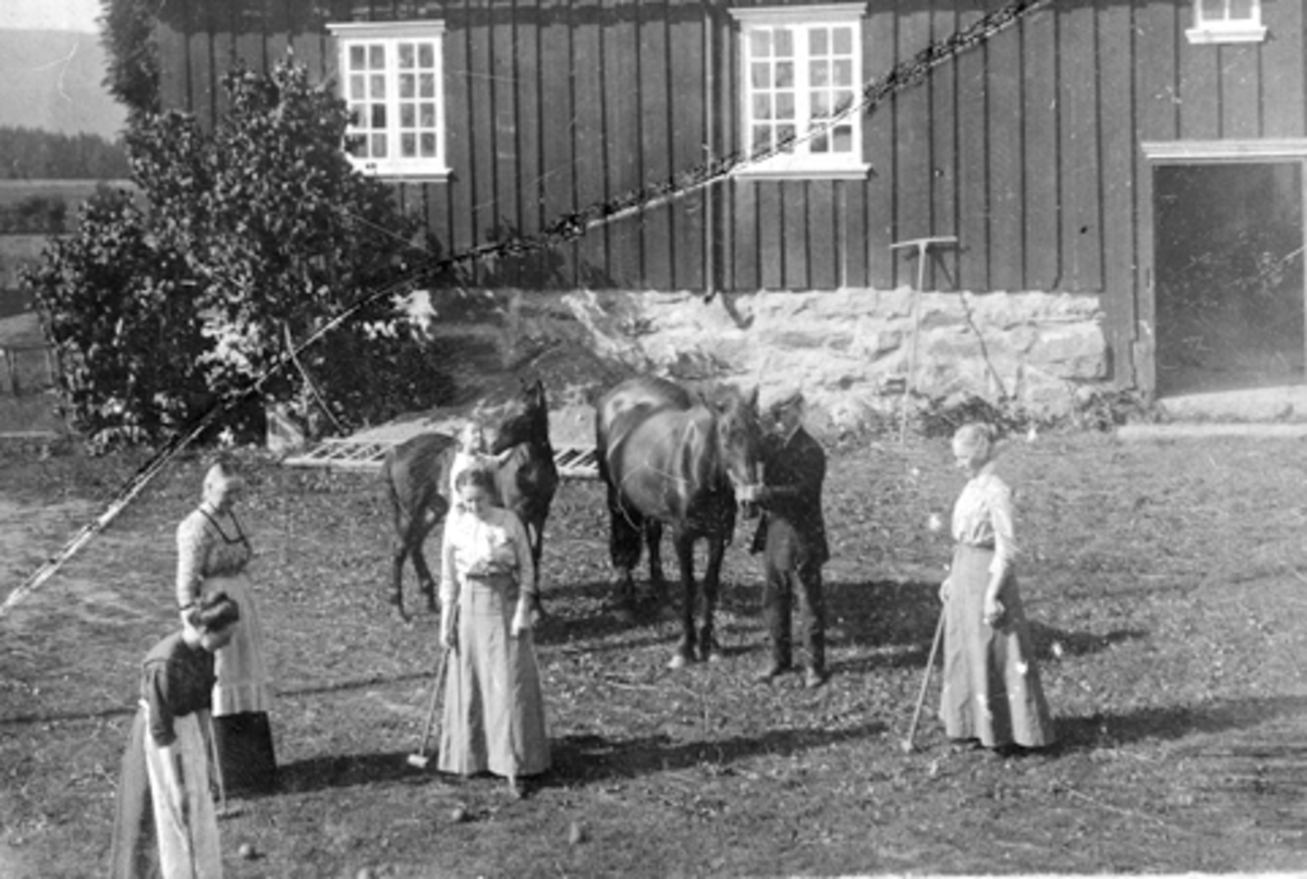 Anno Domkirkeodden, Gruppe kvinner spiller krokket foran drengestua på Mengshoel gard. Mann med to hester på tunet.