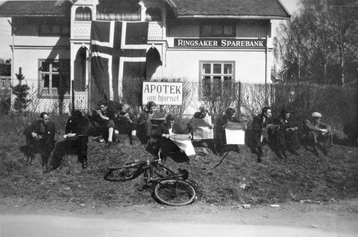Håkon Prestkværn, Moelv, ettermiddagen 8. mai 1945. Oslopressen har nettopp kommet med toget og nyhetene i avisen leses nøye Bildet er tatt utenfor Ringsaker Sparebank. Norsk flagg på bygningen. Fredsdagene.