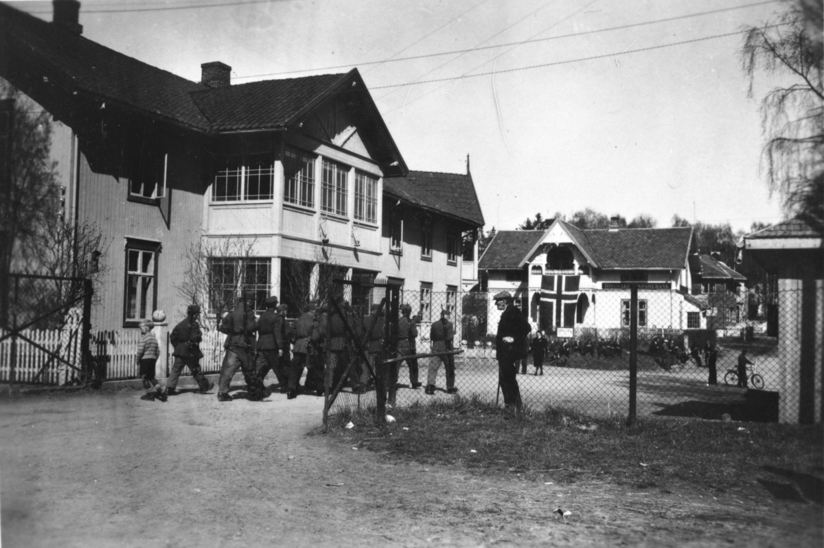 Ukjent, Moelv sentrum, fredsdagene mai 1945. Et skjeldent syn fra maidagene 1945. En tysk tropp marsjerer ut fra jernbanestasjonen fullt bevæpnet.. Det norske flagget henger fra balkongen på Ringsaker Sparebank. Etter kapitulasjonen opptrådte tyskerne disiplinert og ingen episoder inntraff i Moelv.