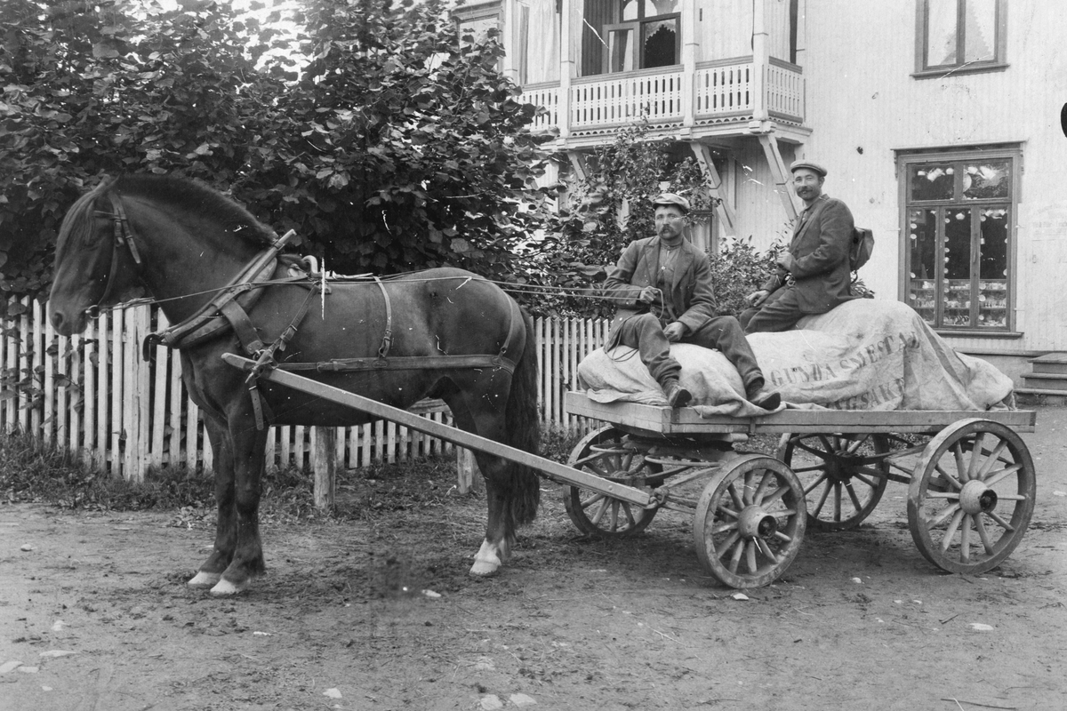 Ukjent, Hest og kjerre, varetransport. Presenning merket "Gunda Smedstad, Ringsaker. " Til venstre Lars "Eriksrud" Mathiasen og ukjent kjører varer for forretningen i Eriksrud innehaver Smedstad. Bildet er tatt utenfor forretningen til Ludv. Larsen som hadde forretning i huset ved Nedre Bruvold (Sundgaard)…
