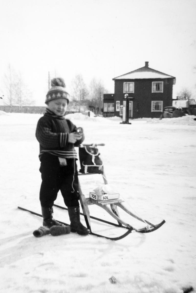 Ukjent, "En liten pjokk fra Ilseng på handletur hos J. P. Kolloen vinteren 1961. Navn ukjent. Huset på andre sida er Kryssby der Andrers og Kjellaug Vestby bodde og hvor det dengang også var kafe. Bensinpumpe foran huset" Barn og spark.