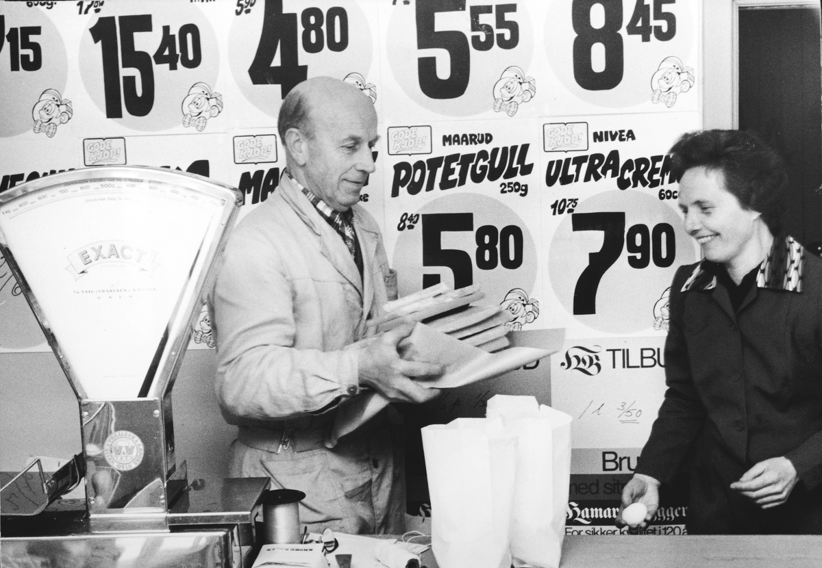 Ukjent, Skogsrud & Haugen Landhandleri på Tangen, 1970-tallet. Butikkvekt, tilbudsplakater. F. v. kjøpmann Per pedersen og ekpeditrise Marit Fagerhaug. Flere opplysninger se boka "Handelssteder i Stange" skrevet av Jan Arne Berg, utgitt av Stange Historielag 2012