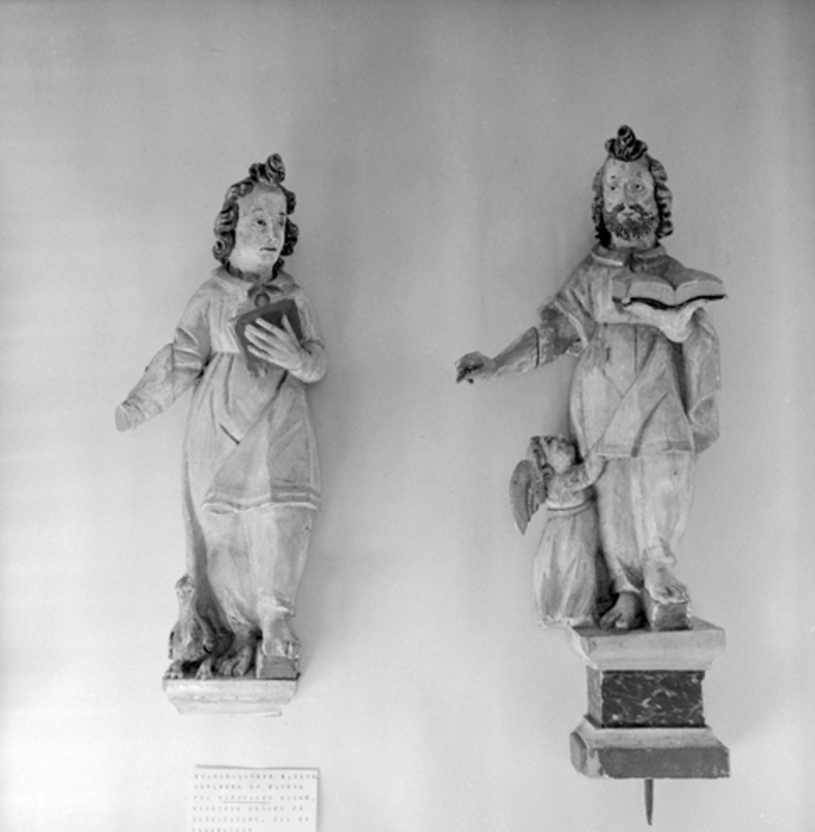 Anno Domkirkeodden, GJENST. KIRKEKUNST, 2 TREFIGURER, RINGSAKER KIRKE. Evangelistene Johannes og Matheus har vært festet til minnetavla over Oluf Thommessøn, men har fra først av hørt med til en prekestol. Brun sokkel med gull og hvitt, brunt hår og skjegg. Hvite drakter med gullborder. Boka t. h. med svart perm, boka…
