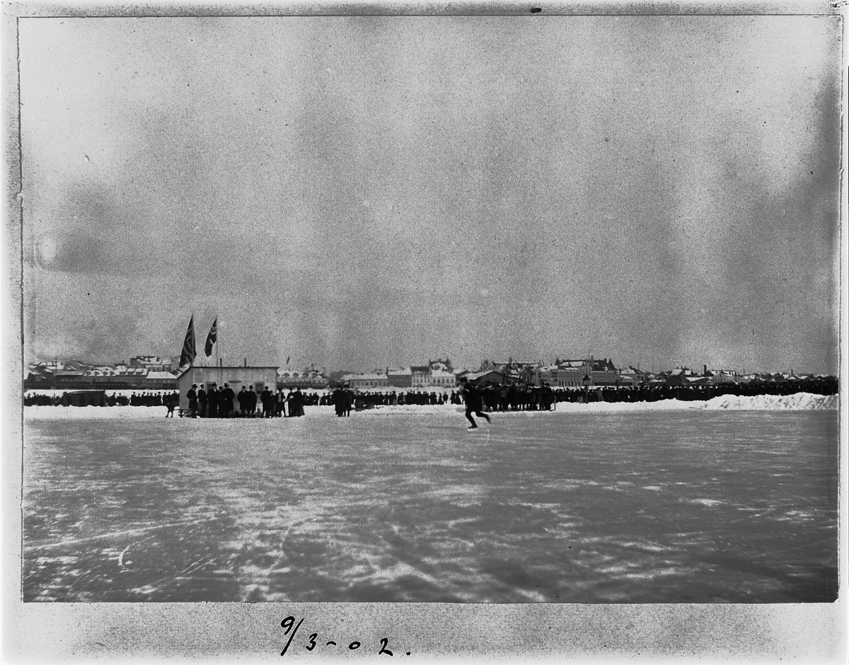 Johan Albert Steen, SKØYTELØP PÅ MJØSISEN, KUNSTLØP, TRIBUNE, PUBLIKUM, FLAGG, 9-3-1902, BEBYGGELSEN I STRANDGATA, HAMAR I BAKGRUNNEN.