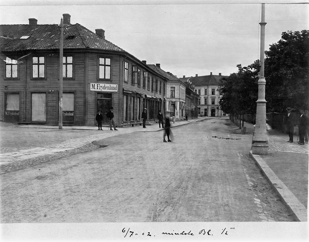 Johan Albert Steen, Hamar, Strandgata, M. Frydenlund, Urmaker Simensens gård, barn og voksne i gata, 6-7-1902. Simensgården t. v. sto ferdig i 1850 og hadde i 2. etg et lokale som fungerte som både kirke og skole (1852-53) i de første årene. Gården brant ned i 1946. I dag ligger "Casa blanca" her.