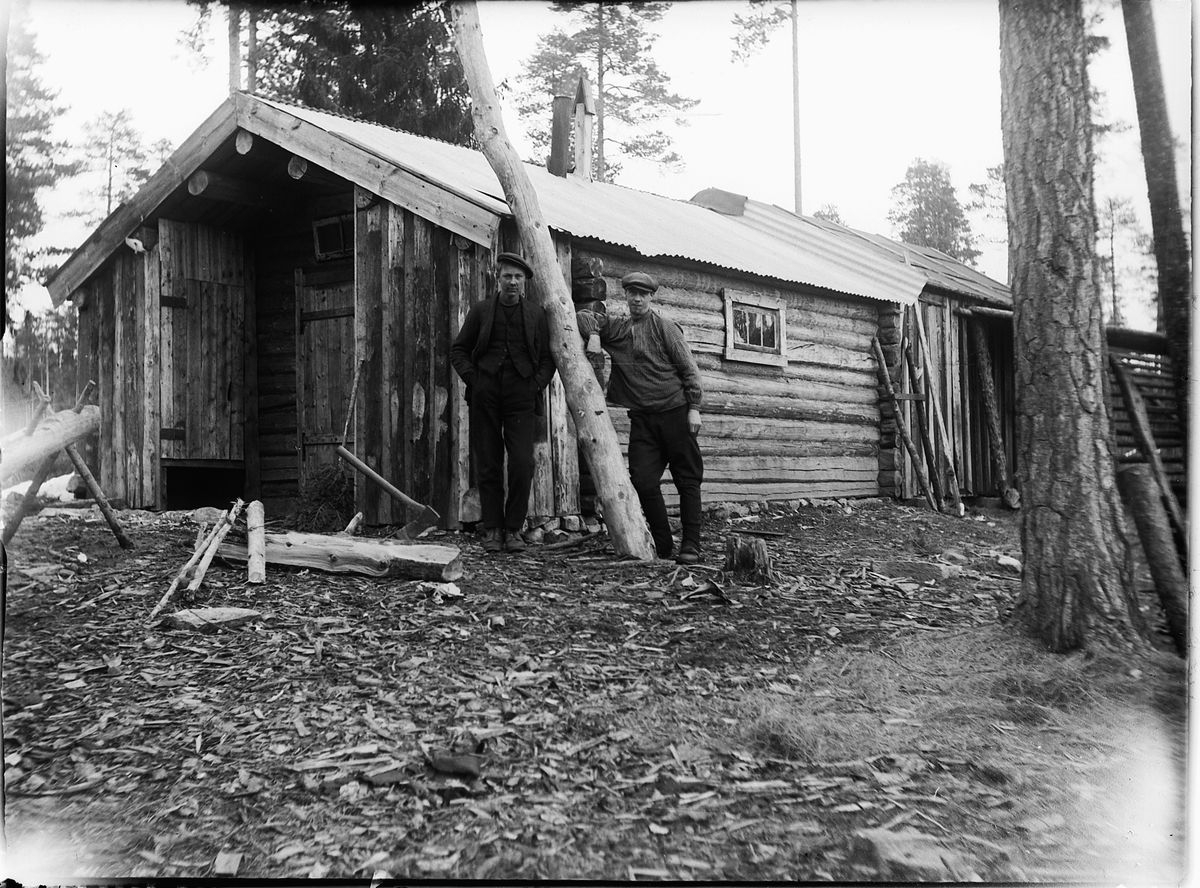 Ole M Martinsen, To ukjente menn i skogen foran ei hytte.