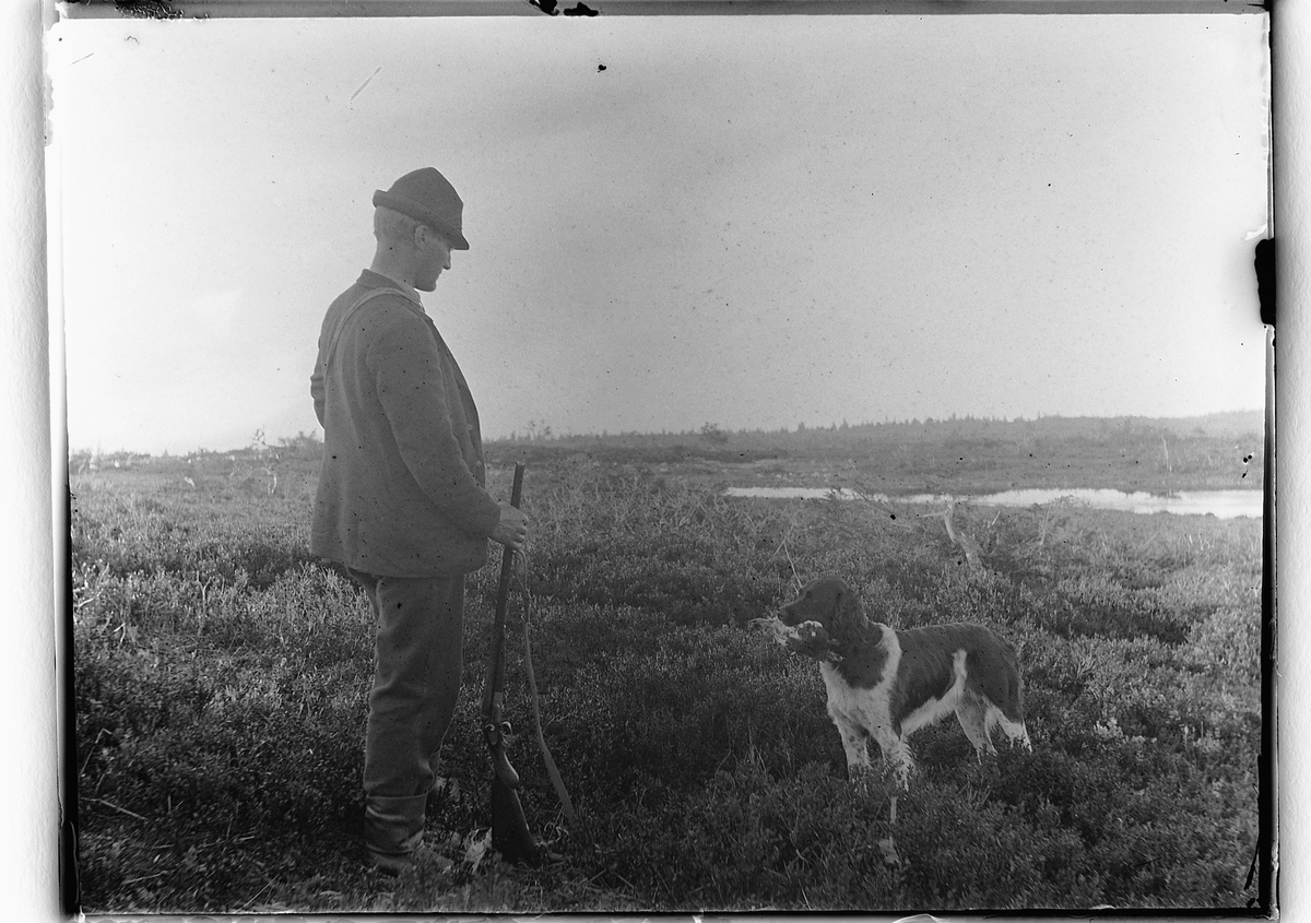 Ole Hansen Løken, FUGLEJAKT, JEGER MED GEVÆR OG FUGLEHUND MED JAKTBYTTE I MUNNEN.