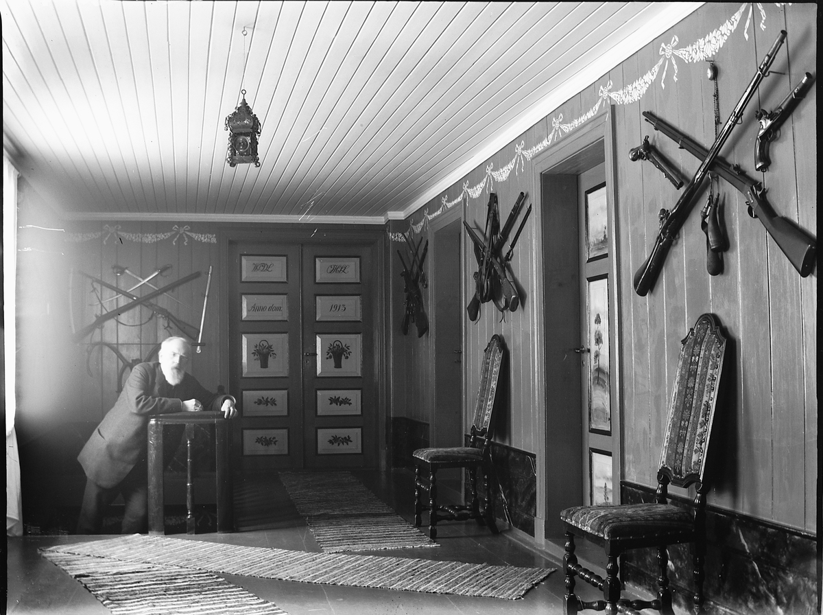 Ole Hansen Løken, LØKEN ØSTRE, VELDRE i Ringsaker, INTERIØR, STUE I 2 ETG HOVEDBYGNINGEN, Wilhelm Peters poserer foran dobbeltdørene inn til salen på Løken østre som han dekorerte og signerte i 1913. VEGGENE PYNTET MED GEVÆR, VÅPEN