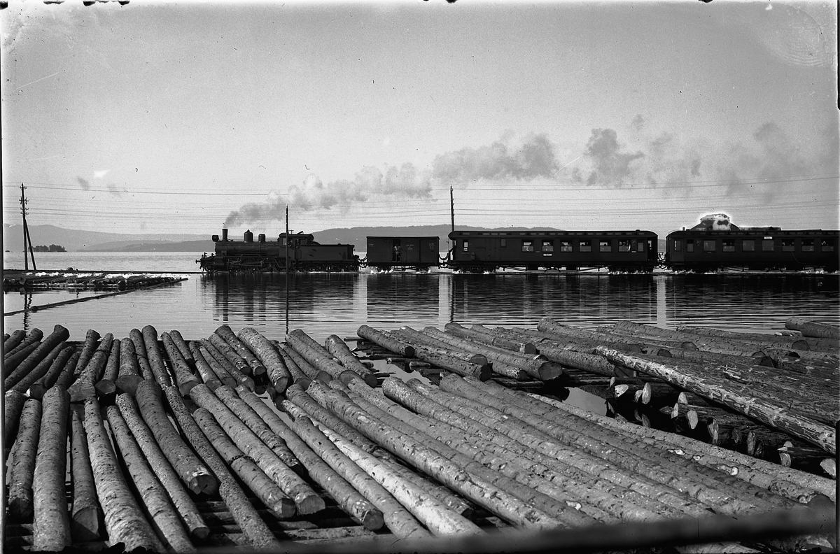 Oscar Løberg, FLOM I MJØSA 1927, TOG OVER ÅKERSVIKA, STANGEBRUA. JERNBANE, DAMPLOKOMOTIV. Vannet sto ei stund enda høyere slik at hjulene gikk halvt under vann. En mann på dressin syklet foran toget for å hjelpe til i tilfelle noe av tømmeret fra lensene fløt over sporene.