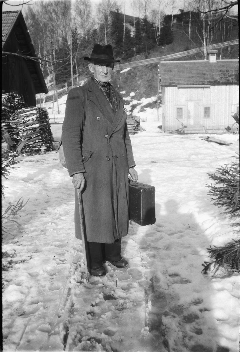 Håkon Prestkværn, Ukjent mann med hatt, stokk og koffert foran bebyggelse.