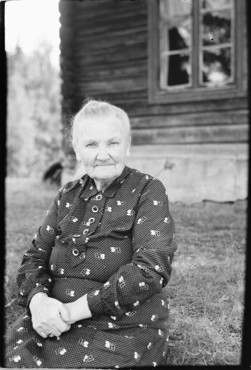 Håkon Prestkværn, Eldre kvinne sitter foran et hus.