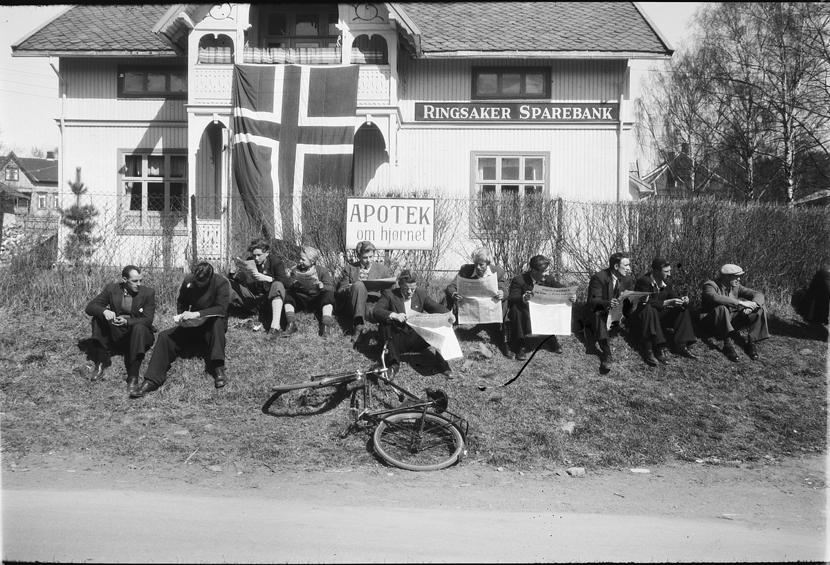 Håkon Prestkværn, Moelv, ettermiddagen 8. mai 1945. Oslopressen har nettopp kommet med toget og nyhetene i avisen leses nøye Bildet er tatt utenfor Ringsaker Sparebank. Norsk flagg på bygningen. Fredsdagene.