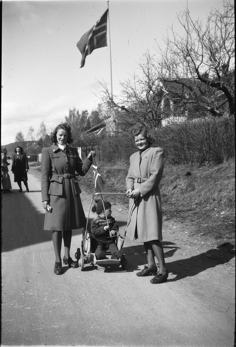 Håkon Prestkværn, To ukjente kvinner med et barn i barnevogn foran det norske flagget.