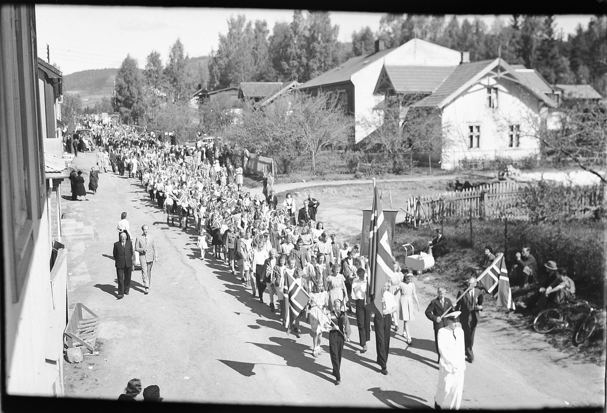 Håkon Prestkværn, 17. -MAI-TOG I MOELV, STORGATA MED HUSET TIL WIEN I BAKGRUNNEN, FORAN LENSMANN BULL I HVIT UNIFORM, NORSKE FLAGG, BARNETOG.