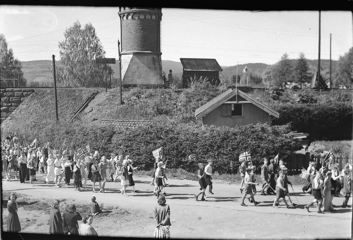Håkon Prestkværn, 17. mai-tog i Storgata, Moelv. Vanntårnet ved Moelv jernbanestasjon i bakgrunnen.