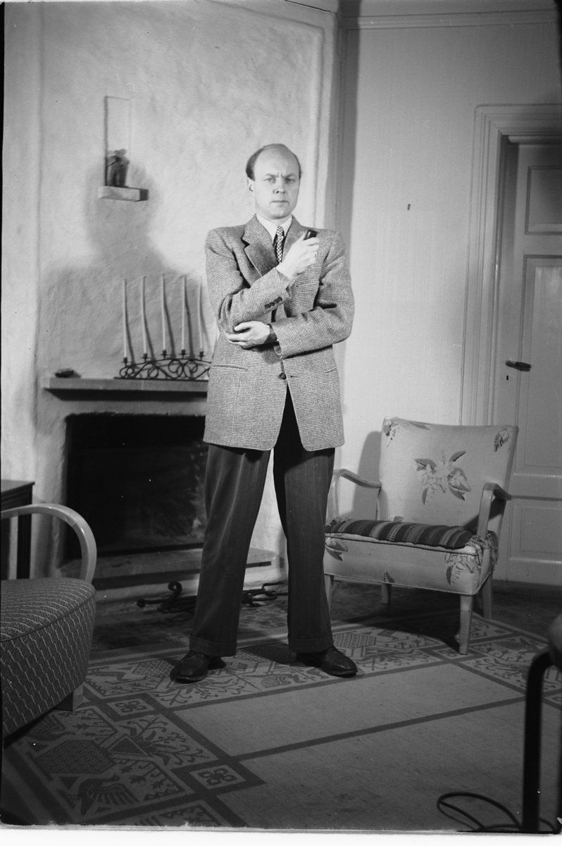 Håkon Prestkværn, Interiør, stue. Mann med pipe står ved en stol og peis.