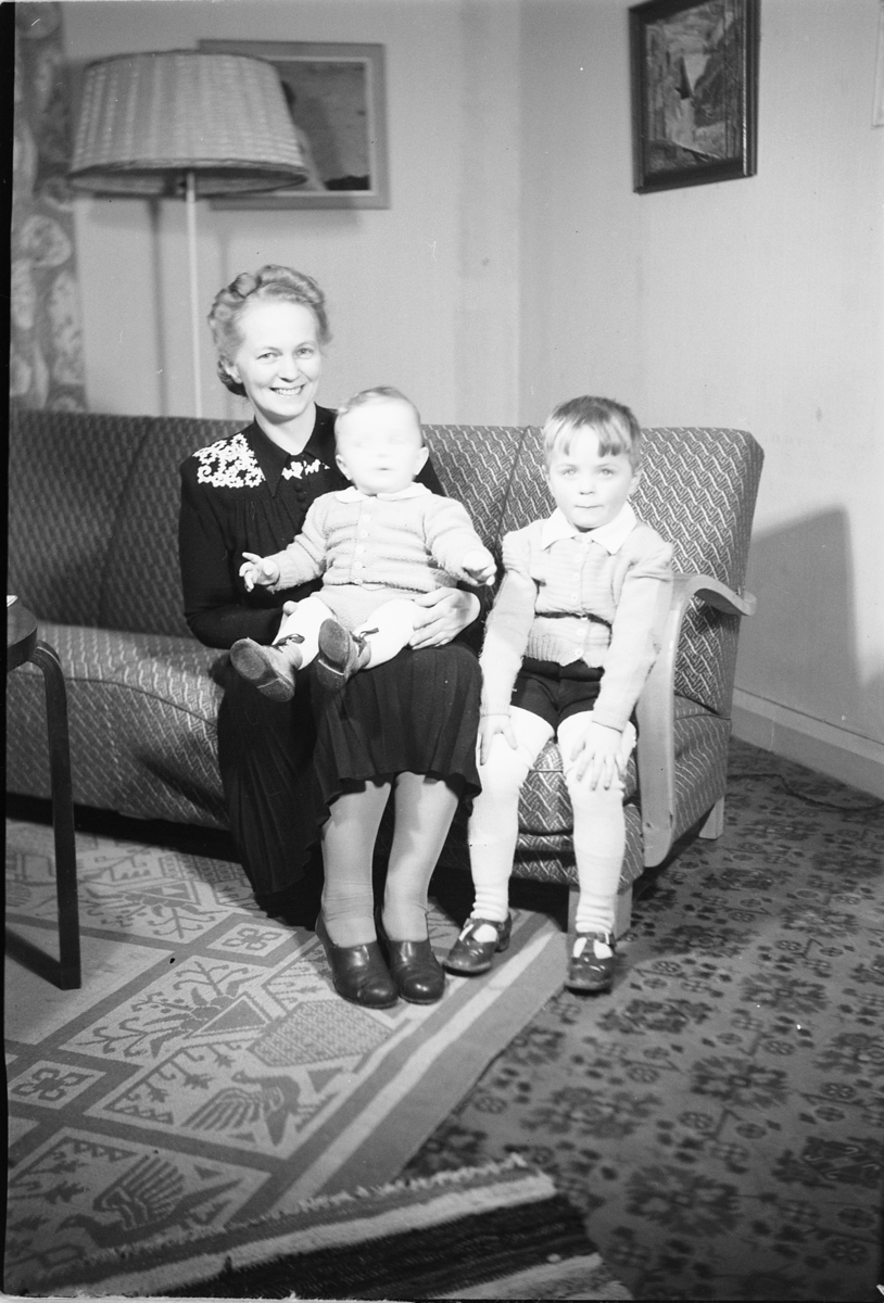 Håkon Prestkværn, Interiør, stue. Eldre kvinne med to barn i en sofa.