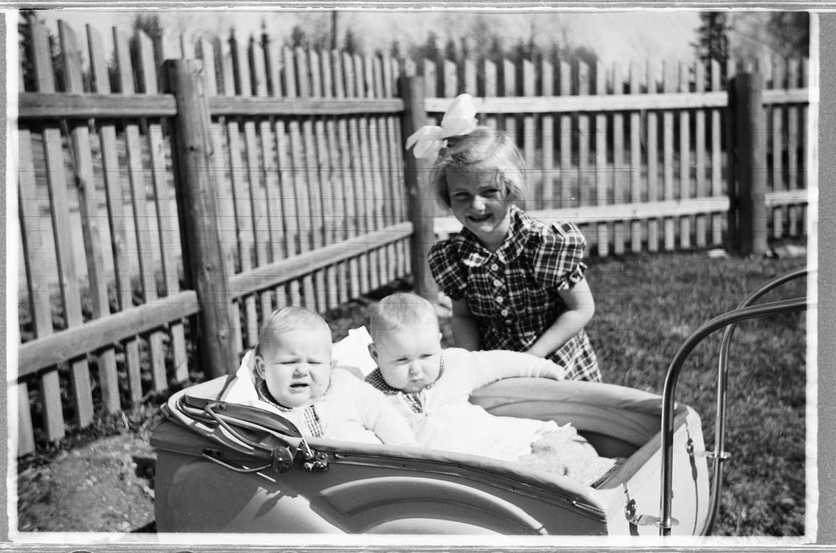 Håkon Prestkværn, To babyer i en bagnevogn og ei jente med sløyfe i håret. Tvillingene i vogna er Knut og Bjørg Svenskerud f. 1949. Jenta med sløyfe er Inger Svenskerud f. 1945. Bildet er tatt ved Åsmarka kirkestue.