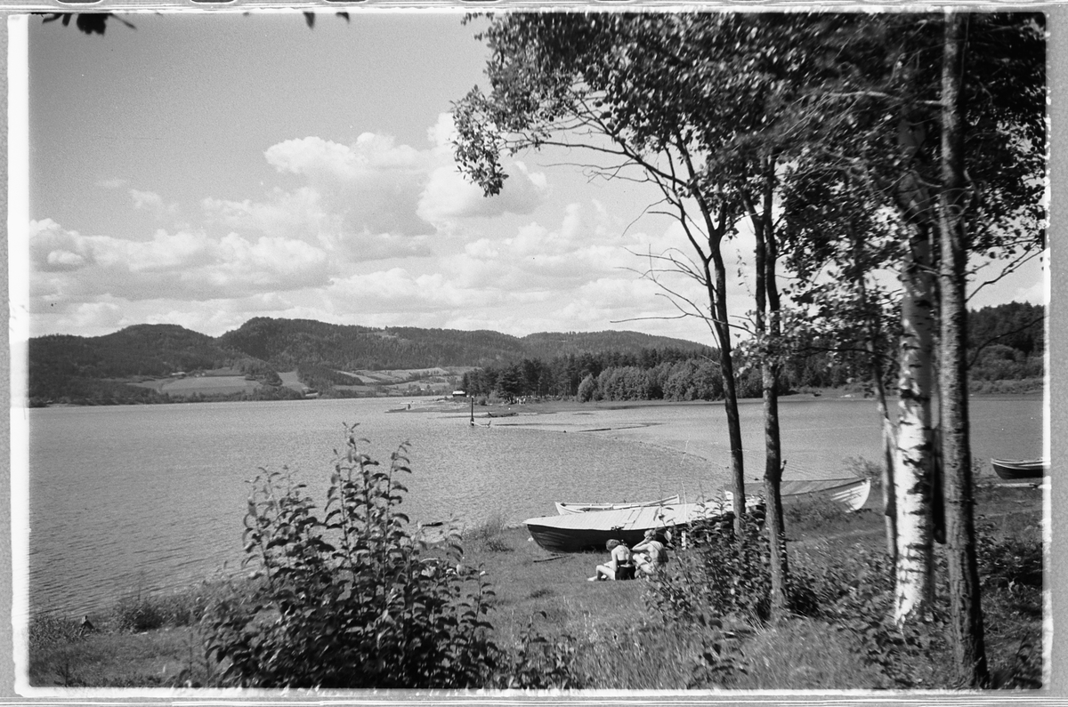 Håkon Prestkværn, To kvinner sitter i gresset ved vannkanten og robåter. Mjøsa ved Moelv.