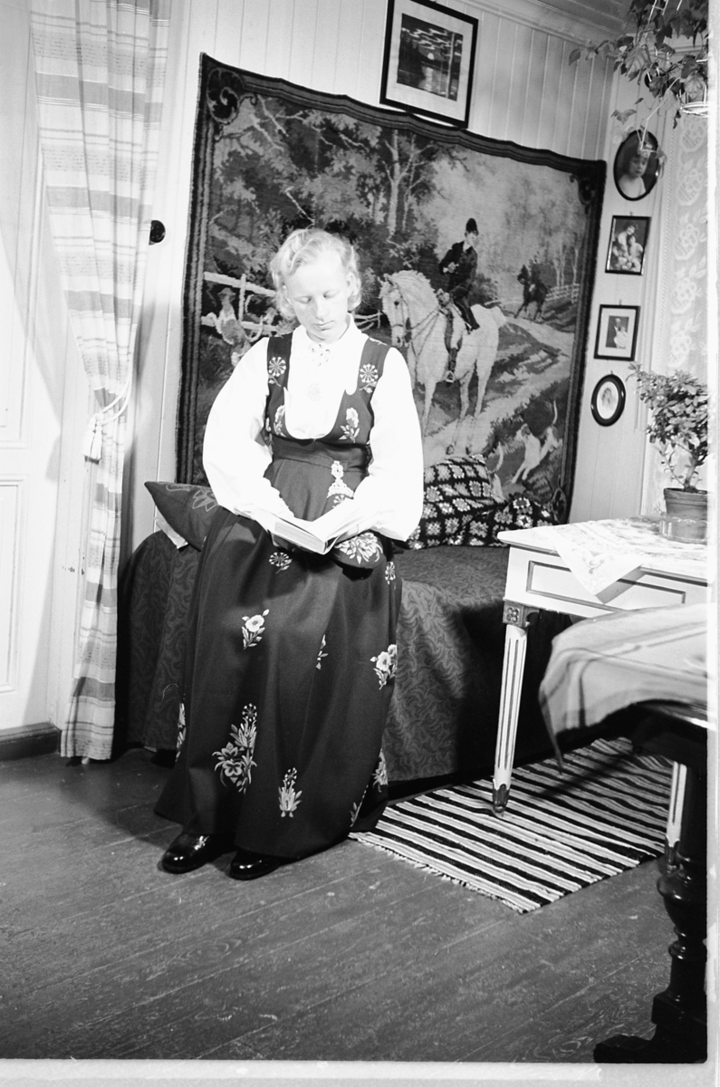 Håkon Prestkværn, Interiør, stue. Kvinne i bunad sitter i en sofa og leser ei bok.