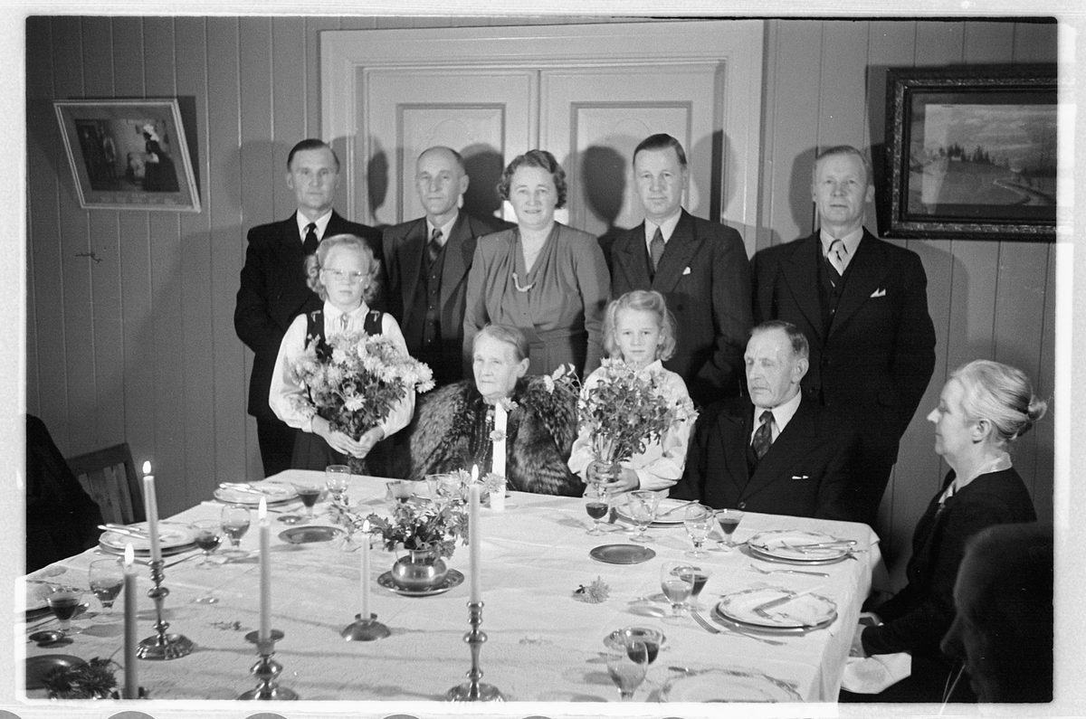 Håkon Prestkværn, Interiør, stue. Familiegruppe samlet ved et dekket bord.