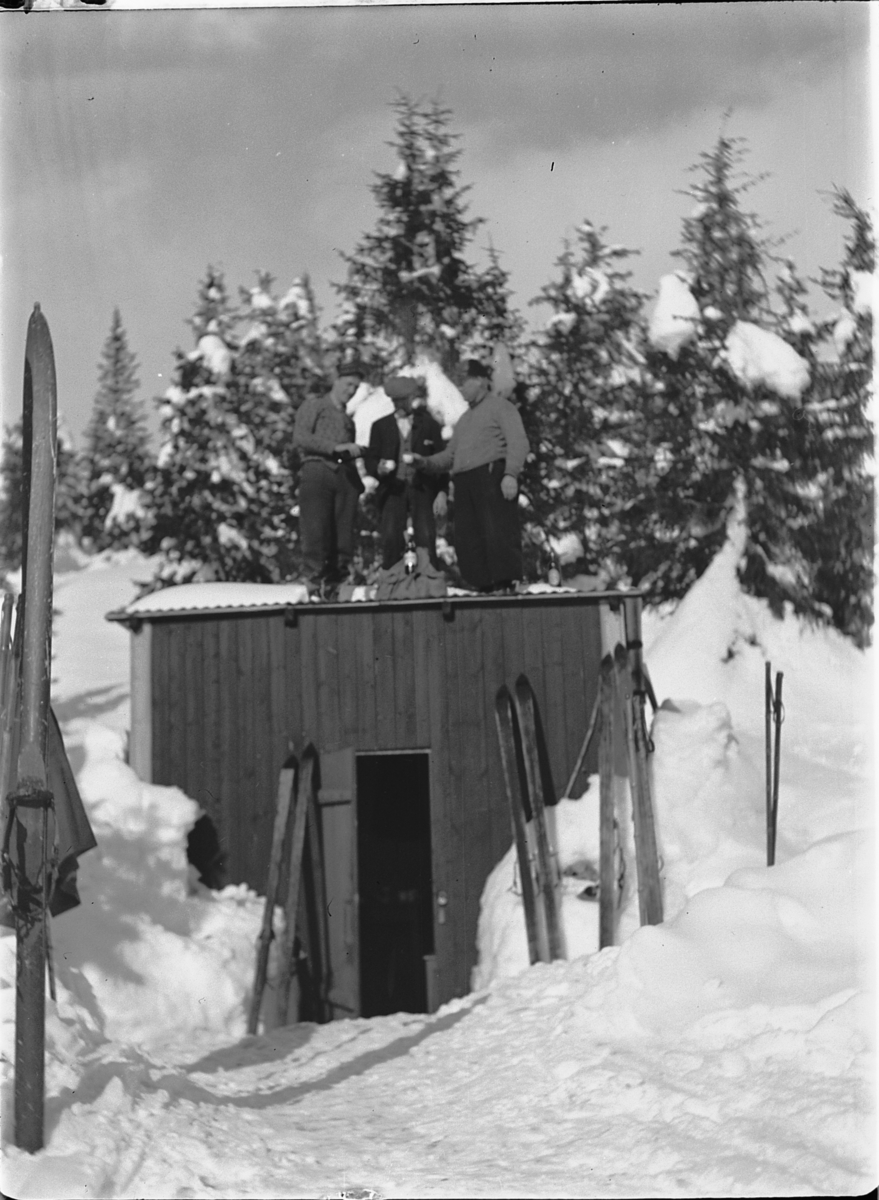 Ole M Martinsen, Tre menn på skitur tar seg litt drikke på taket av ei hytte.