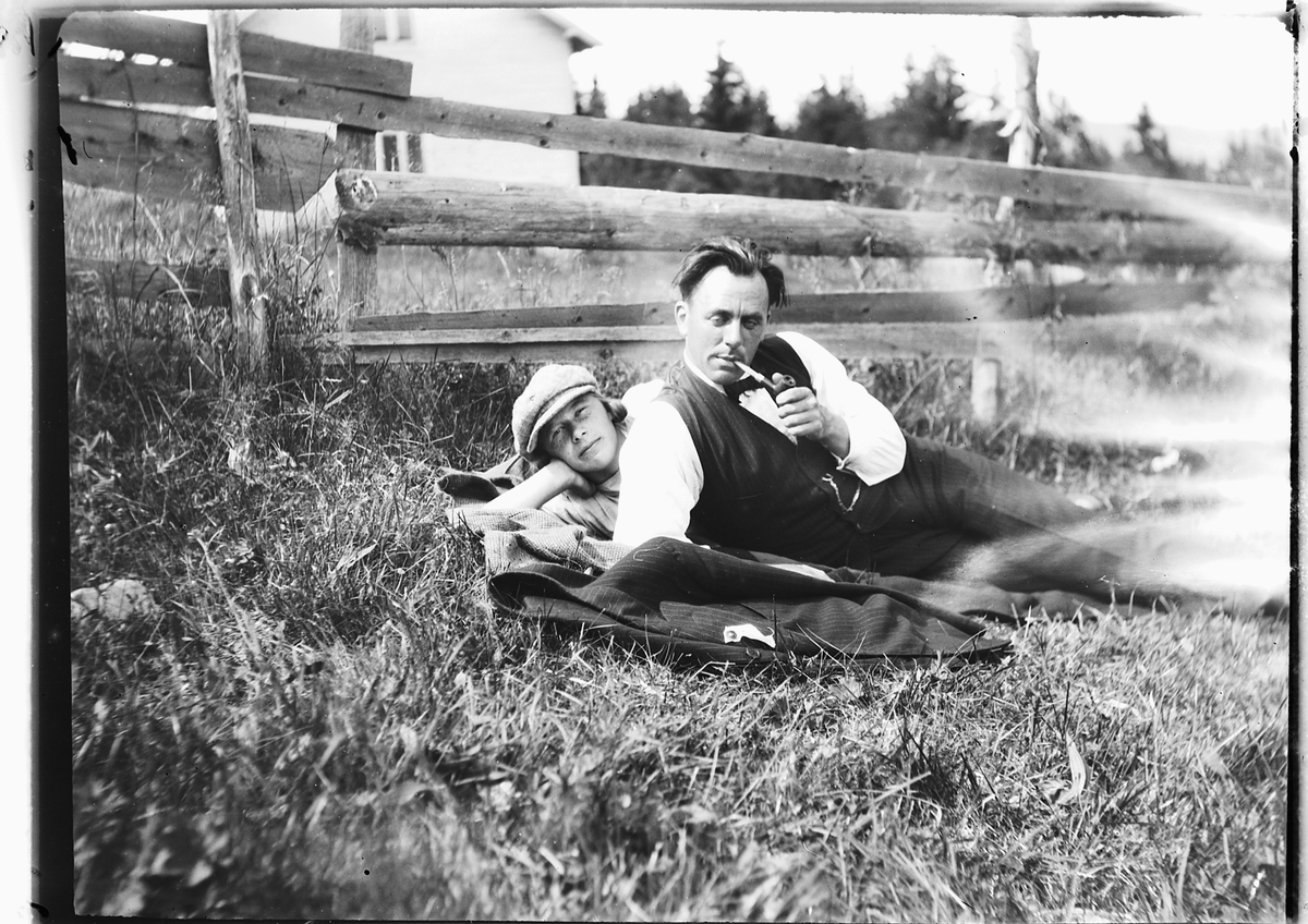 Ole M Martinsen, Ukjent kvinne, og mann som røyker pipe, ligger i gresset ved en skigard.