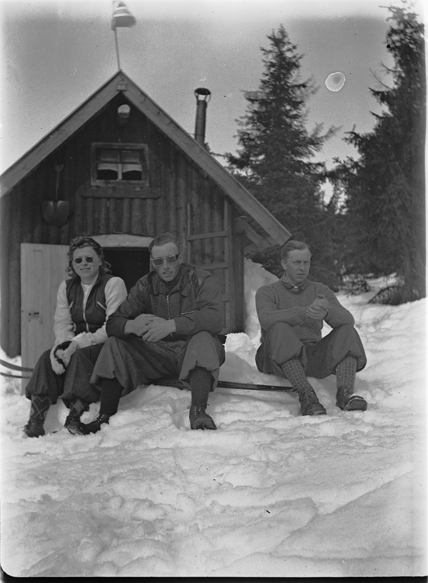 Ole M Martinsen, Ukjent kvinne og to menn utenfor ei HYTTE i skogen.