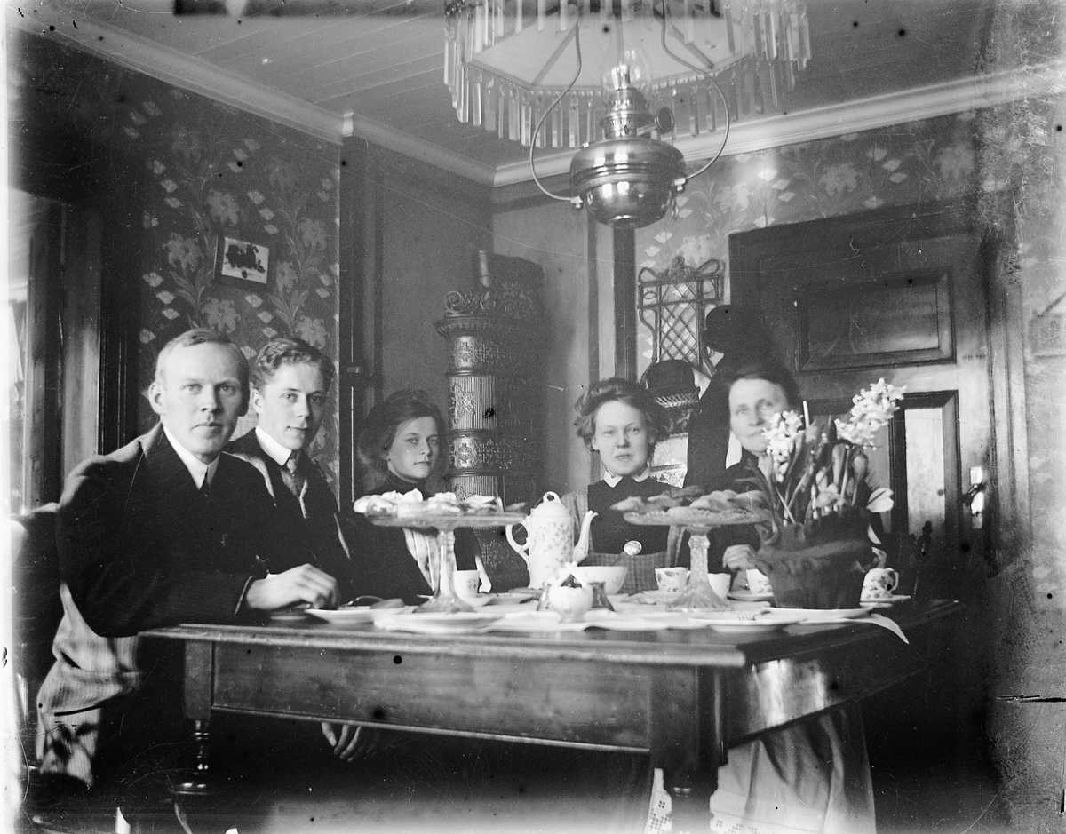 Johs. (Johannes) Johannessen, Fra høyre: Fru Lovise Johannessen, Netta (tjenestepike), Anna Marie Johannessen, nn og Otto? Interiør, stue. Familien Johannessen drikker kaffe ved stuebordet, parafinlampe, kaffeservise.