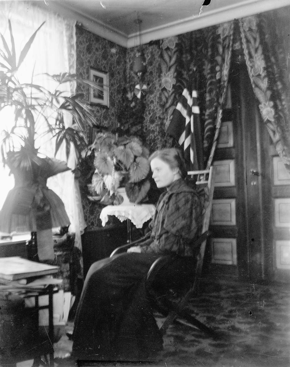 Johs. (Johannes) Johannessen, Interiør, stue. Anna Marie Johannessen, datter til bakemester Olaus og Lovise Johannessen, i gyngestol foran det norske flagget.