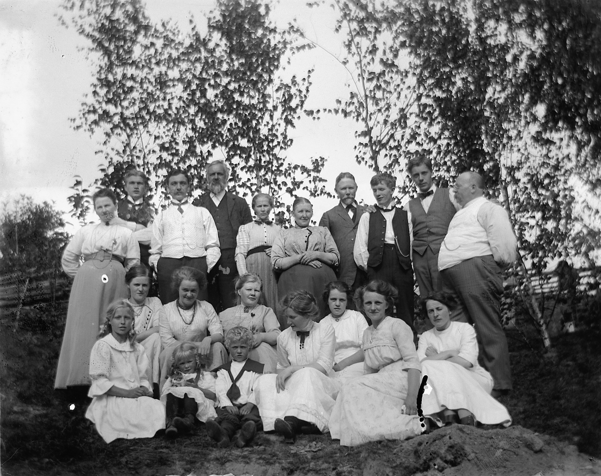 Johs. (Johannes) Johannessen, Stor gruppe kvinner, menn og barn. Stående bak til venstre: Lovise Johannessen. Stående bak til høyre: Olaus Johannessen. Ole Maurud,nn, Paul Maurud, Ingeborg Maurud (søster til Lovise). Sittende fra høyre: Louise Johannessen. Sittende nr 3 fra venstre med stort mørkt kjede er Aagoth Morbech. Sitte…