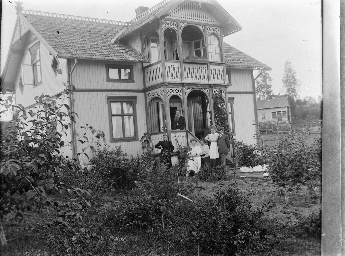 Ragnvald Andersen, Gr. 6. Familien Danielsen. Fokstuenhuset. To menn og fire kvinner foran veranda. Sveitserhus med hage. Landhandler og jernbanesmed Oluf Danielsen, Lundberg, (Haraldstad), Furnesvegen i Hamar. Fotografens svigerfamilie. Eksteriør.