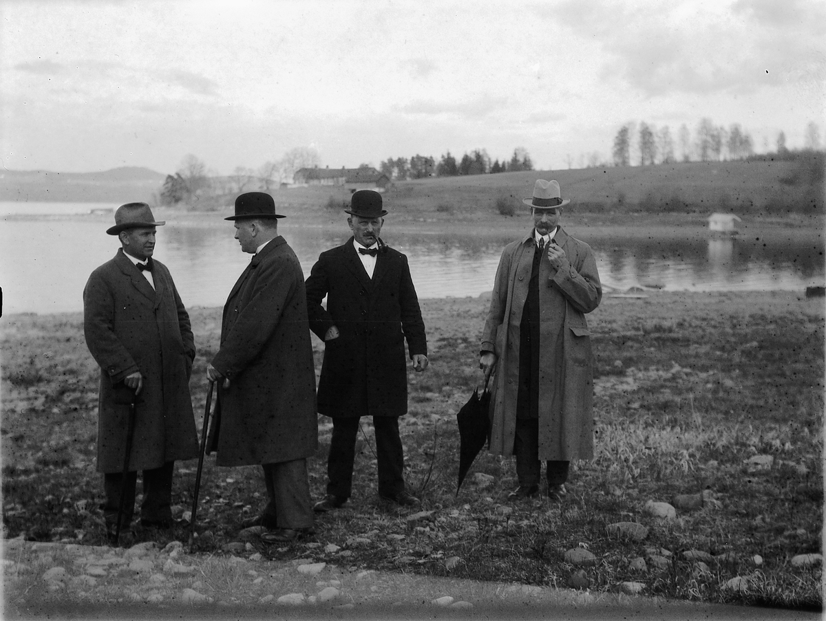 Ragnvald Andersen, Fire menn i hatt, spaserstokk og paraply ved vannkanten.
