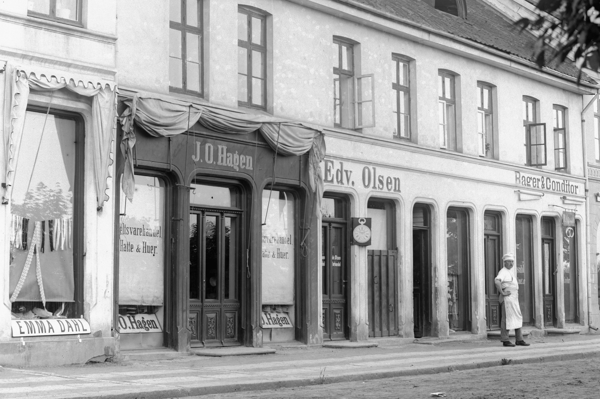 Hans Emil Erichsen, Strandgata 41-43 i Hamar. Forretningene til Emma Dahl, J.O. Hagen, Bager & Conditor Edv. Olsen, J.O. Lier og søstrene Lier. Personene på bildet er bakermester B. Opsahl.