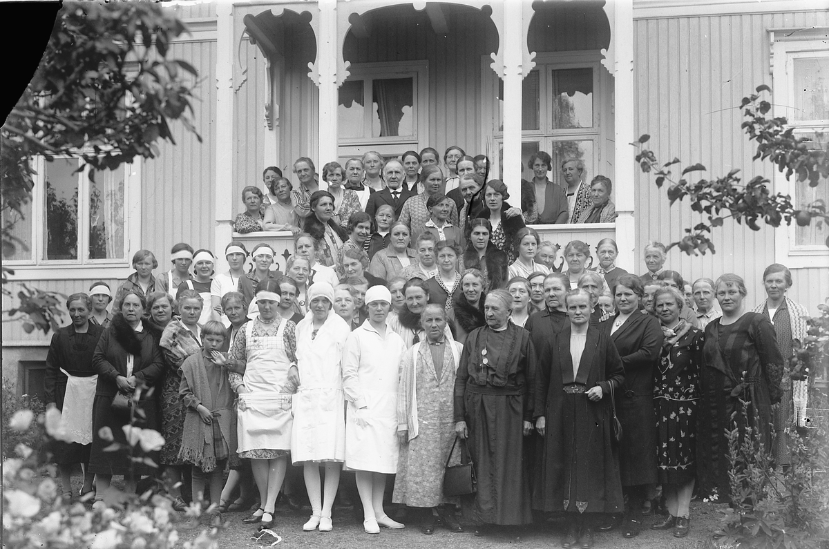 Oscar Løberg, Medlemmer av Døves Vel, Hamar, samlet på en veranda. På verandaen nr. 4 f. v. (inntil søylen) fru byggmester A. Johansen, i midten (eneste mann) Hansen. Bak han t. v. fru urmaker Ødegaard. Til høyre øverst i trappa bestyrerinne frøken Heiberg, med hånda på skulderen til kassereren: fru banksjef Ell…